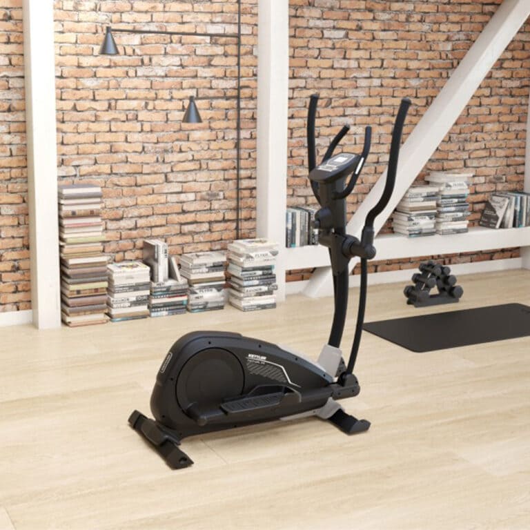 Dviratis treniruoklis patalpinamas patalpoje su medinėmis grindimis ir mūrinėmis sienomis. Prie sienų sukrautos knygos, o fone ant kilimėlio matyti nedidelių svarmenų rinkinys.